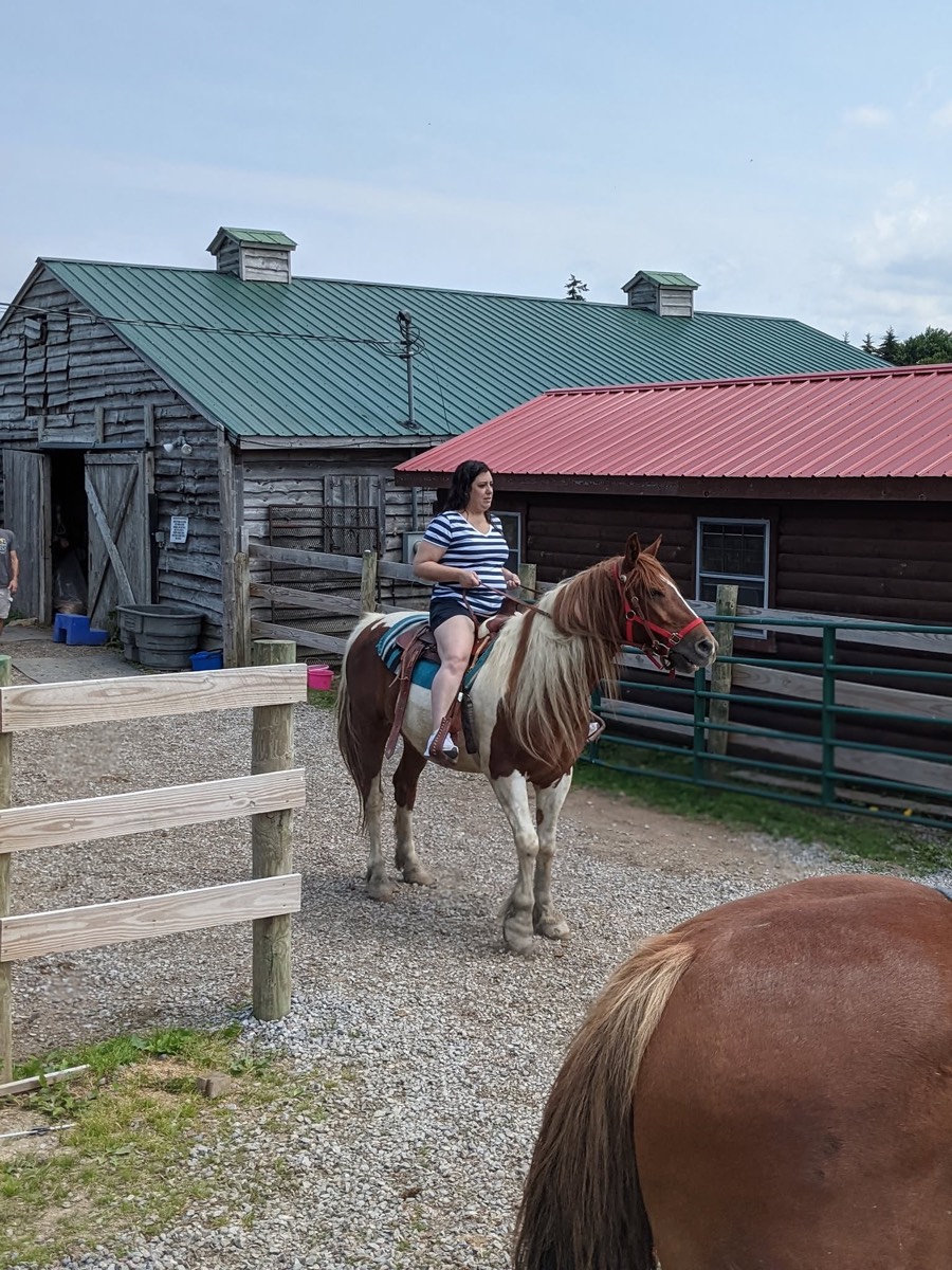 A woman in a navy and white striped top mounted on a brown and white paint horse on the gravel, a weathered grey barn with a green metal roof behind her and a red-roofed log building beyond.