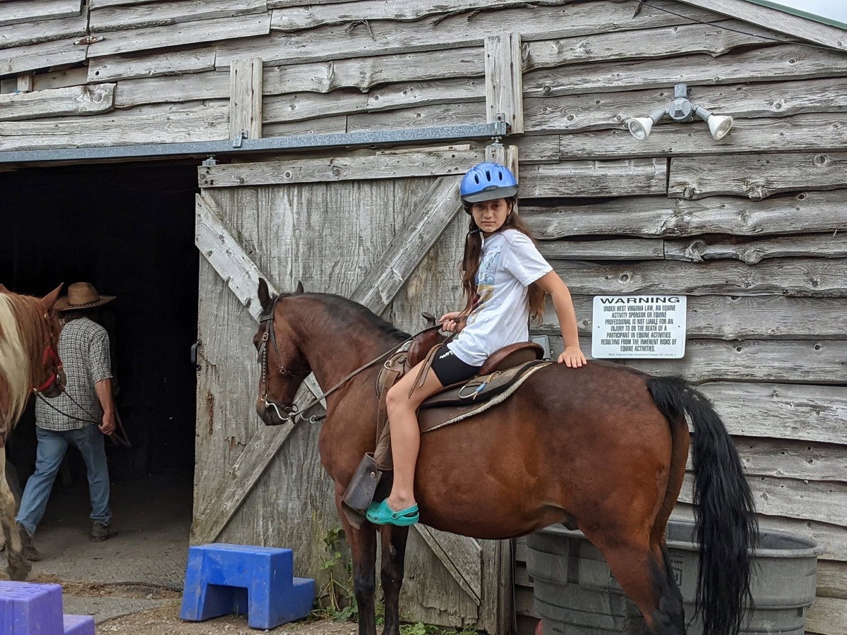 A girl in a blue helmet sitting on a bay horse beside the open door of a gray barn, purple plastic mounting steps on the gravel below.