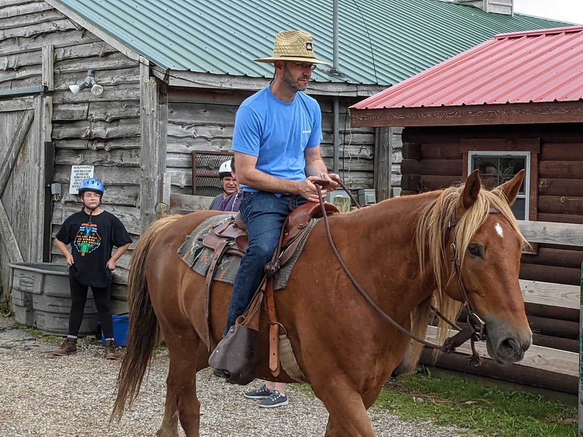 A man in a straw hat and blue shirt riding a chestnut horse past a weathered barn, a girl in a blue riding helmet watching from the shade and a second adult rider in a white helmet waiting behind him.