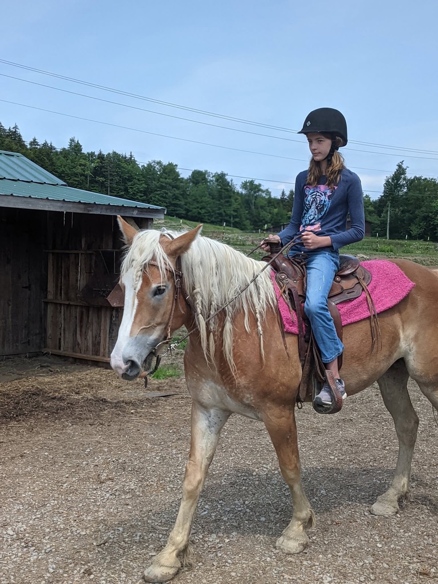 The same palomino walking along a gravel track with a young rider aboard, barn and green ridge behind.