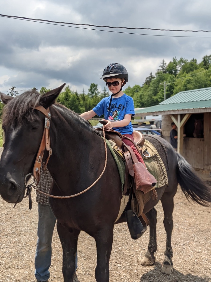 A child in a helmet and sunglasses mounted on a dark horse, waiting on the gravel with the barn roofline behind.