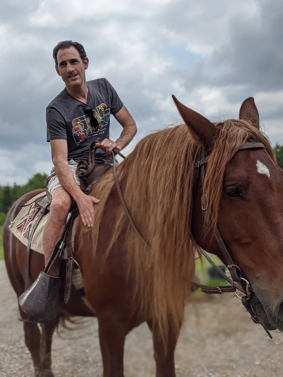 A man on horseback beside a second chestnut horse, both facing the camera on a gravel track under cloud.