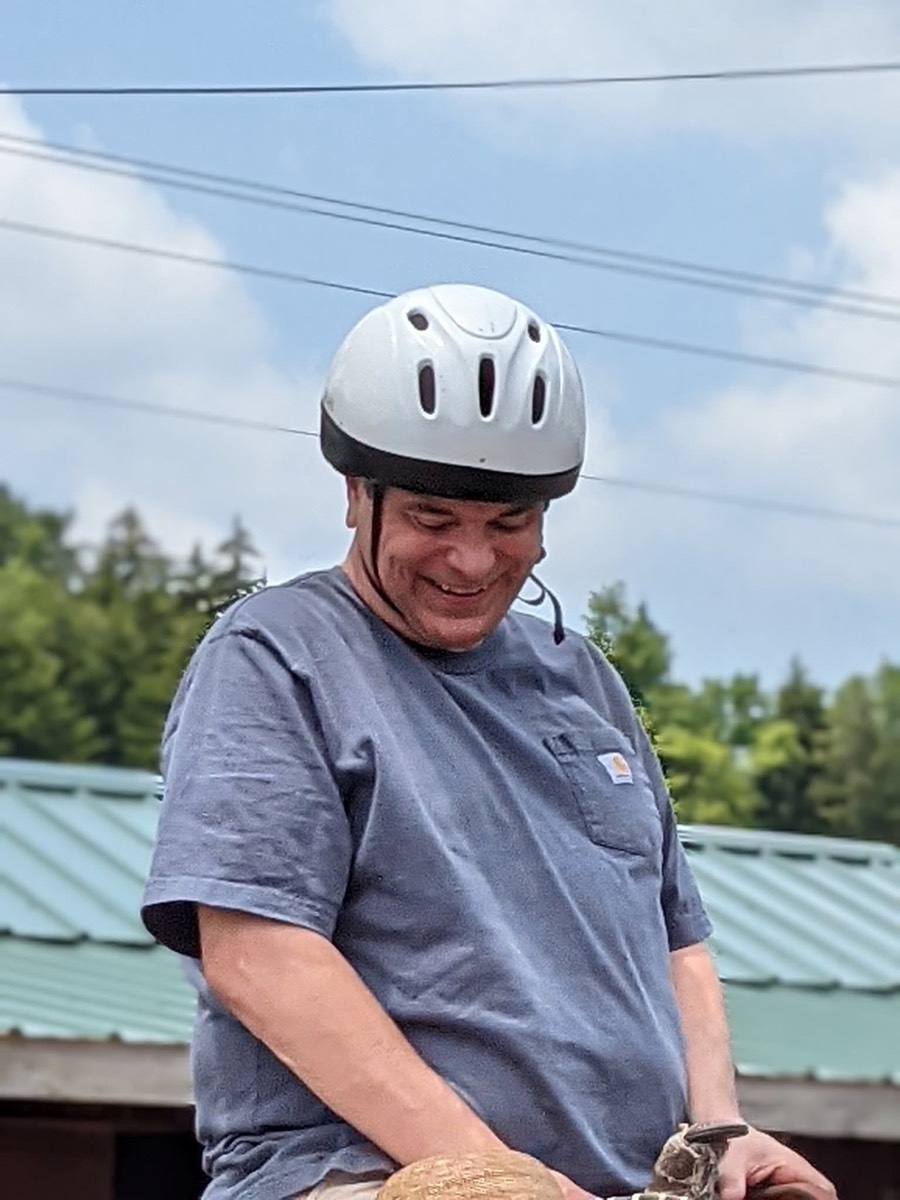 A man in a white riding helmet looking down and smiling, a horse's mane just visible at the bottom of the frame.