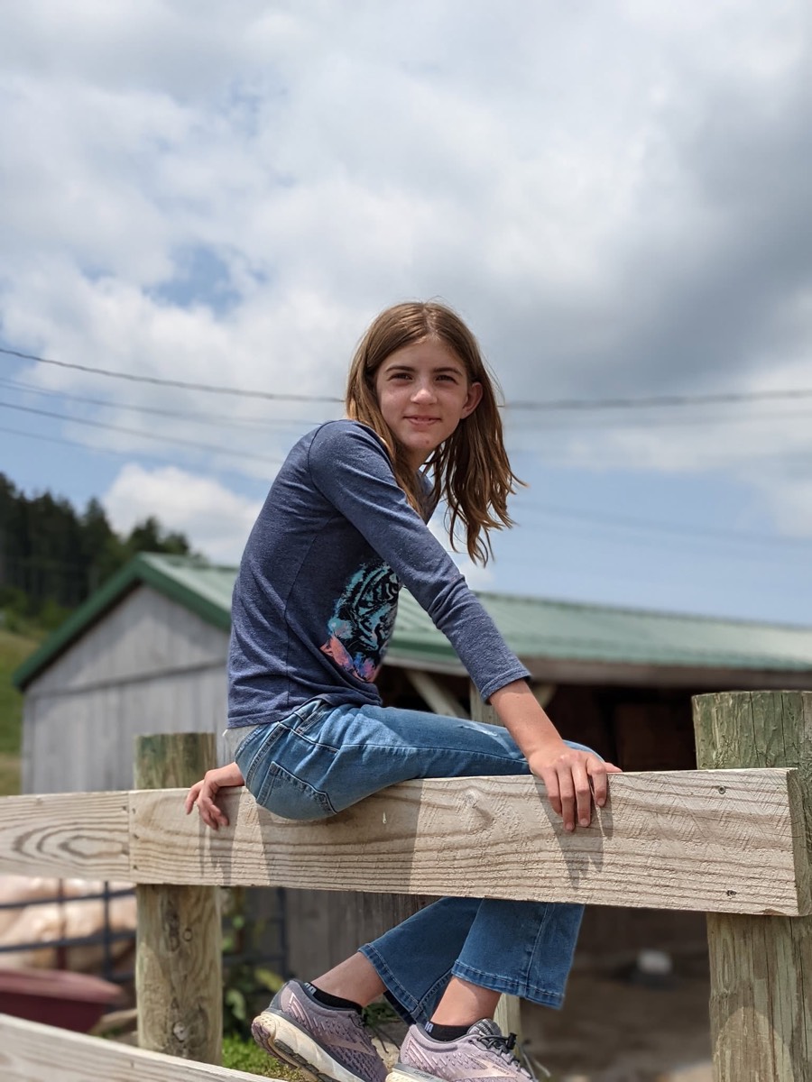 A girl sitting up on a wooden fence rail in front of the barn, arms braced, smiling at the camera.