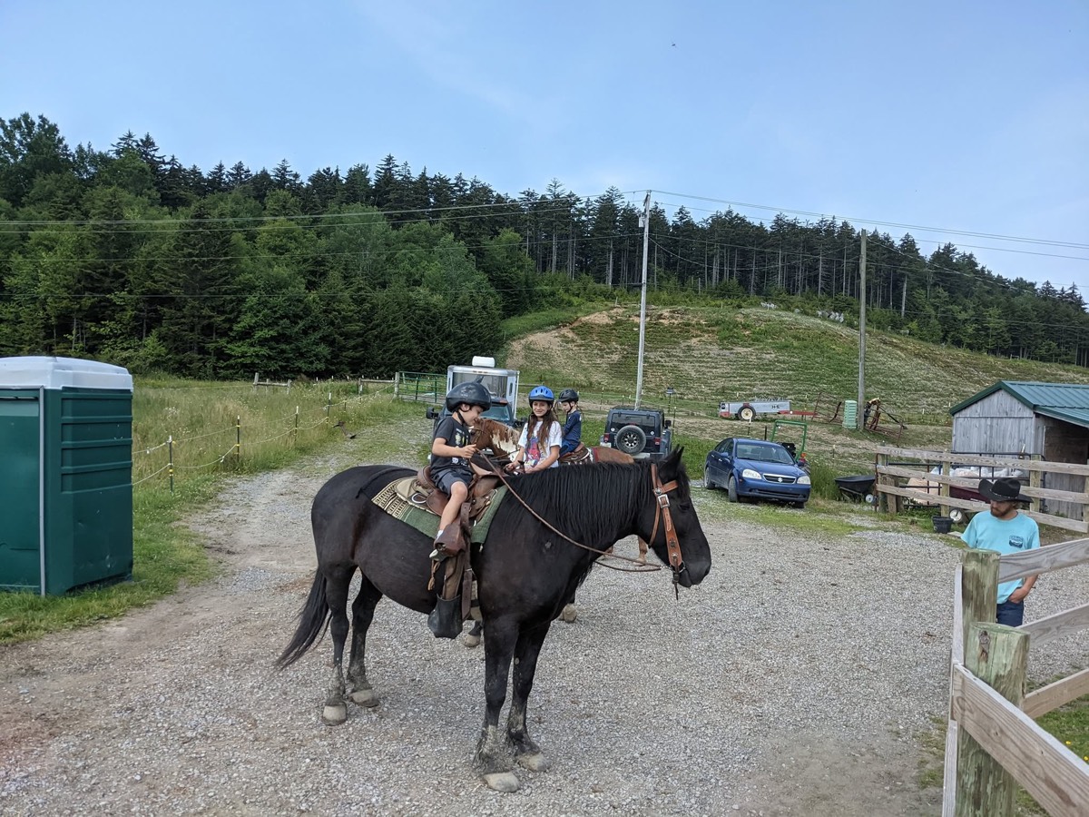 Riders on horseback setting off along a gravel track past a portable toilet and parked cars, open grass slope beyond.