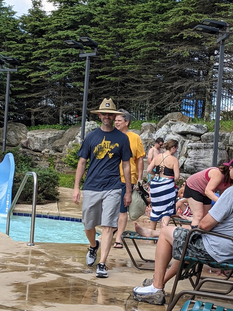 People walking and sitting around a mountain-top swimming pool with lounge chairs, rocks and spruce trees behind.