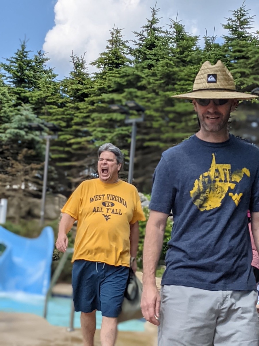 Two men walking along the poolside, one in the gold West Virginia shirt mid-shout, the other in a sun hat and gray shirt.