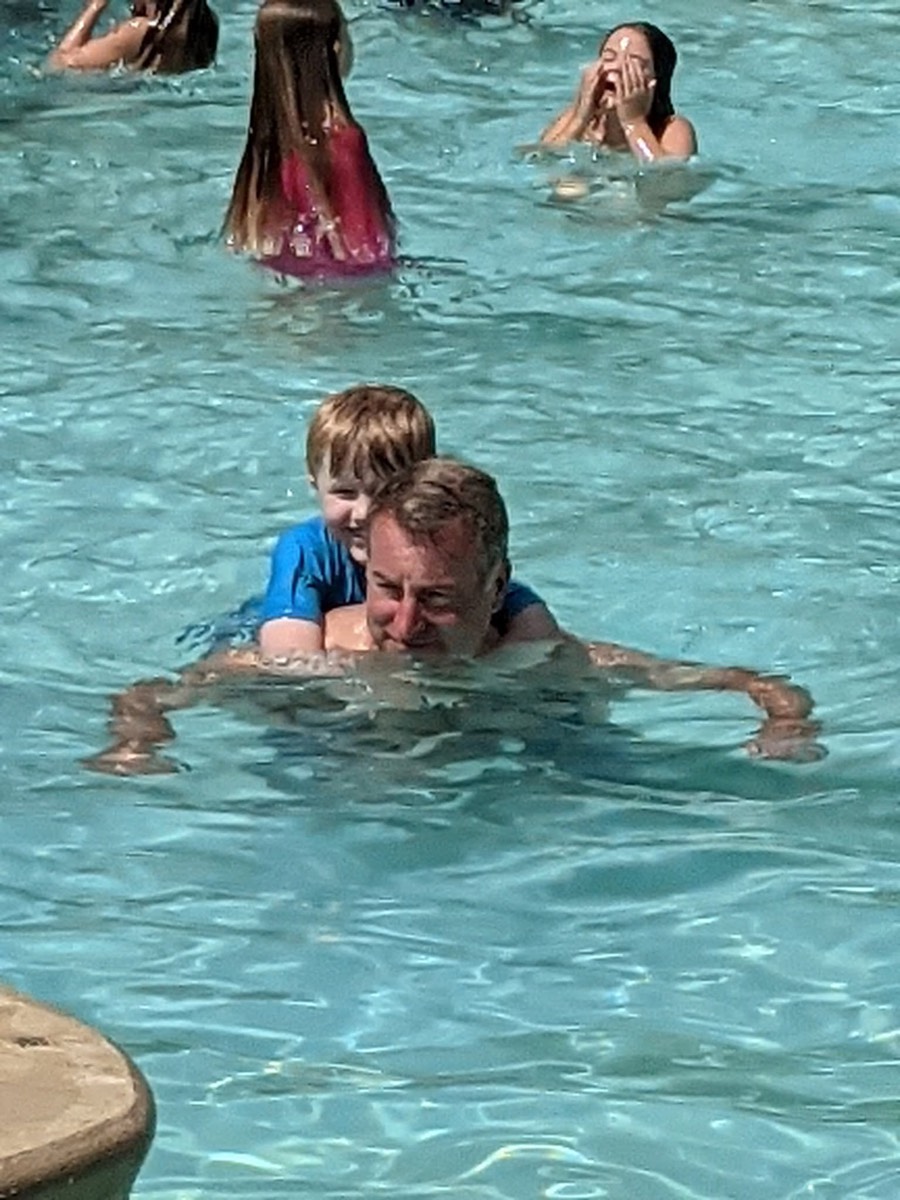 A man in the pool with a small boy on his back, both grinning, other swimmers behind them.