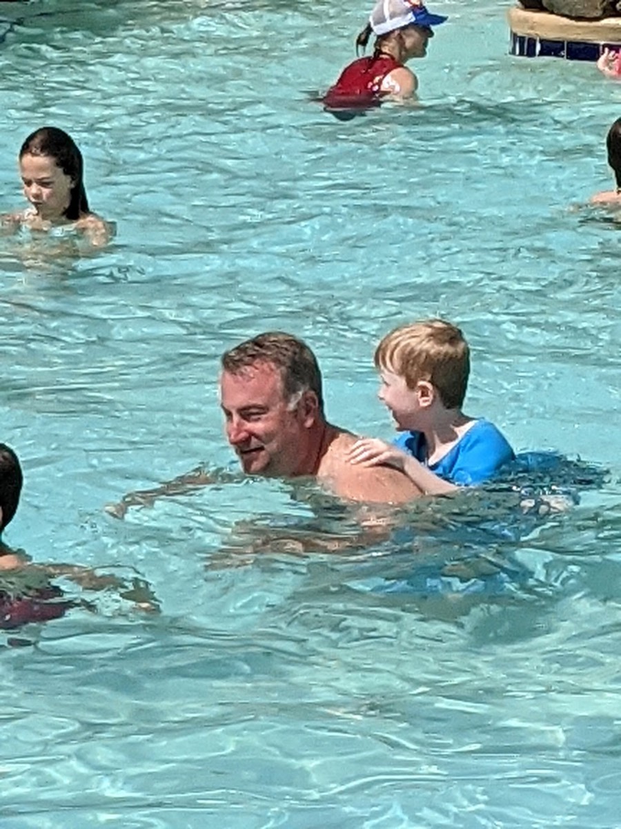 The same man and boy in the pool, the boy's arms round his neck, laughing, other children in the water behind.