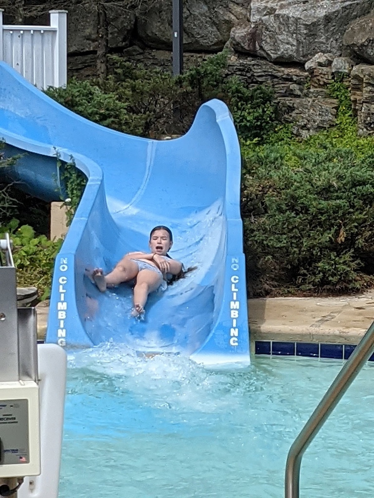 A child coming out of the bottom of a curling blue waterslide into the pool in a burst of spray, rocks and shrubs behind.