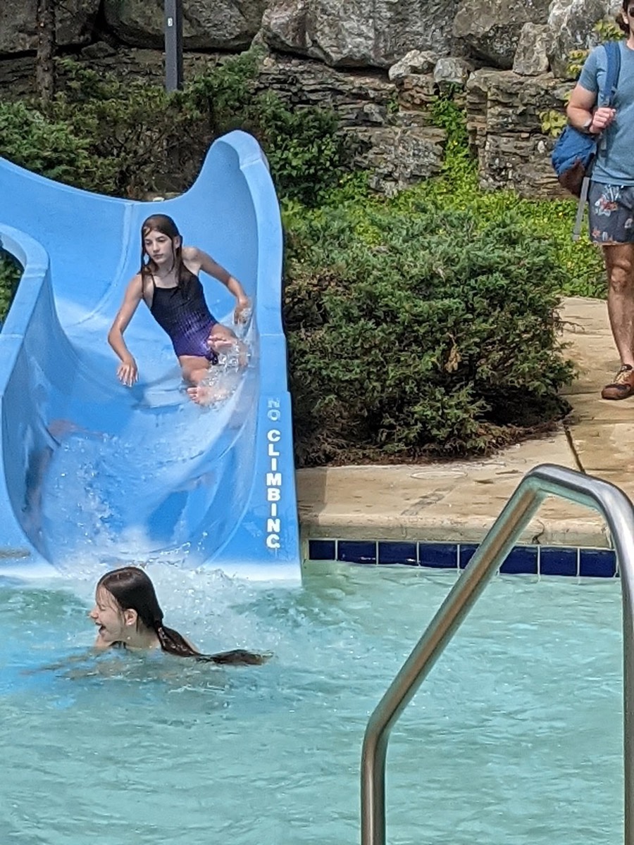A girl standing in the mouth of the blue waterslide as another child surfaces in the water below.