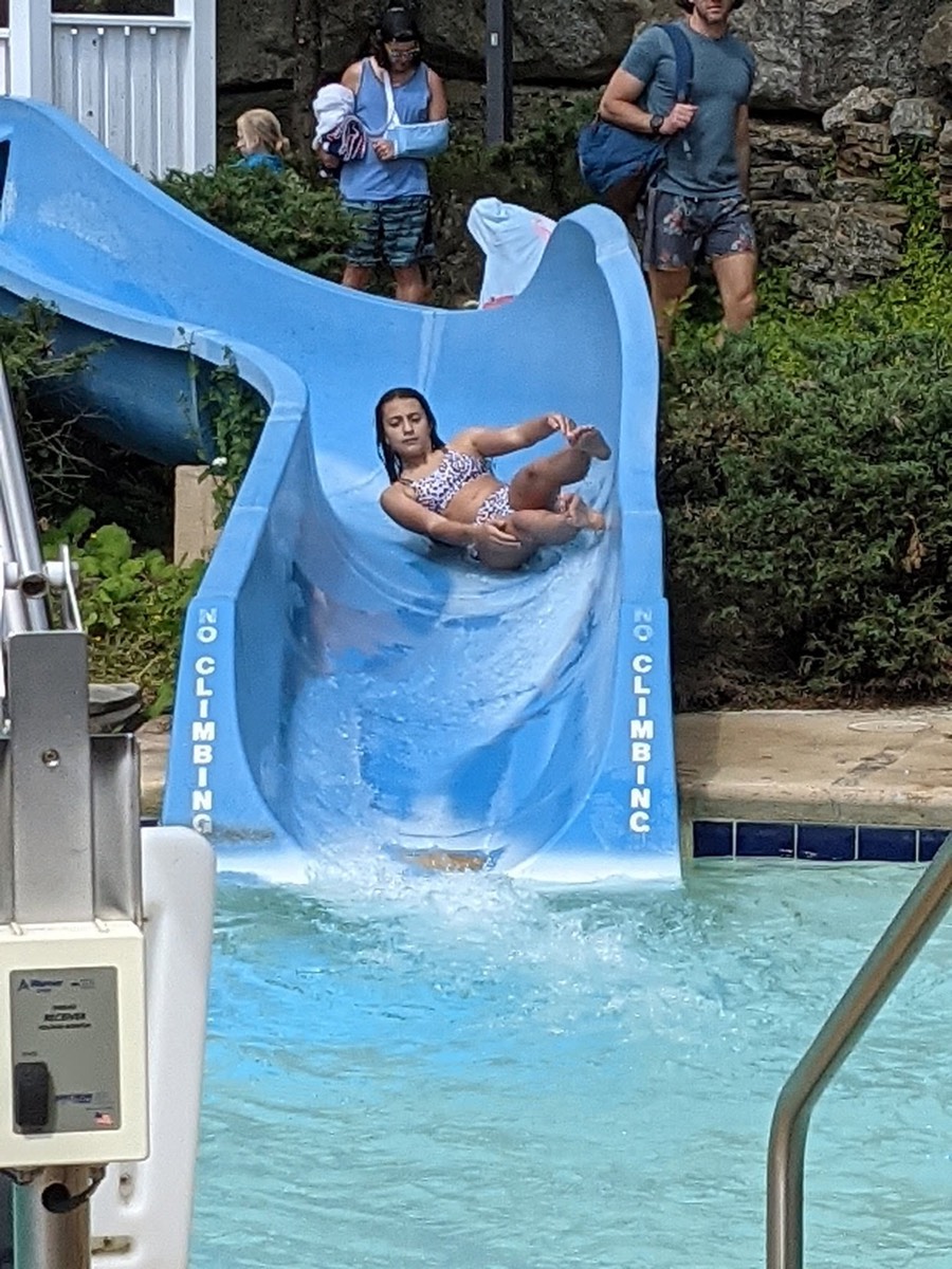 A child lying back in the curve of the blue waterslide, arms wide, coming down toward the pool.