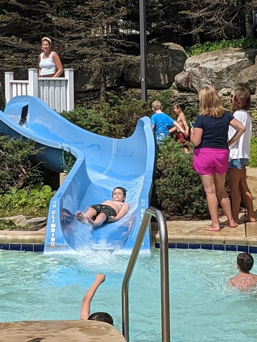 A child sliding down the blue waterslide into the pool, watched by others standing at the poolside.