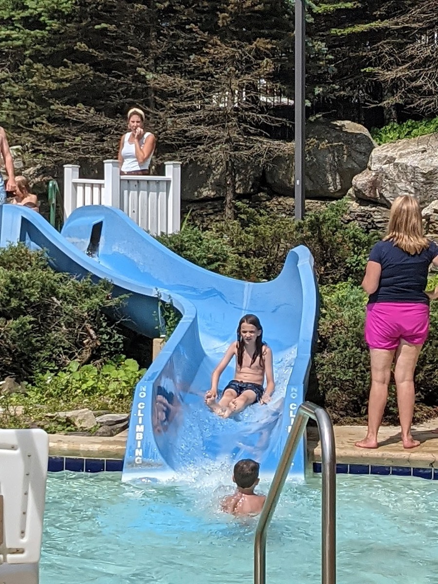 A girl coming down the blue waterslide toward the pool with people watching from the poolside.