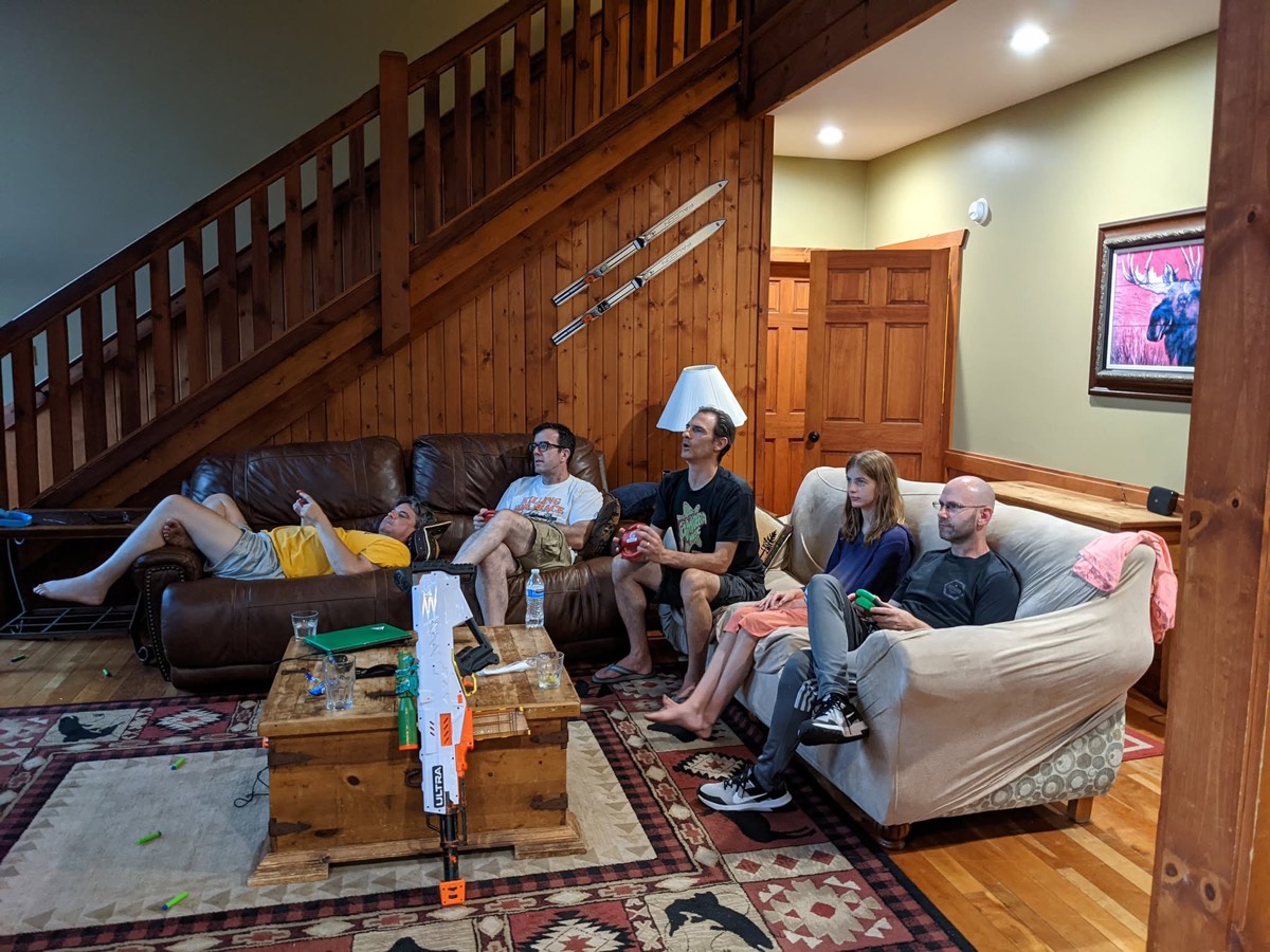 A cabin living room with a wooden staircase, several adults sprawled across sofas and armchairs, a low table covered in drinks and games.