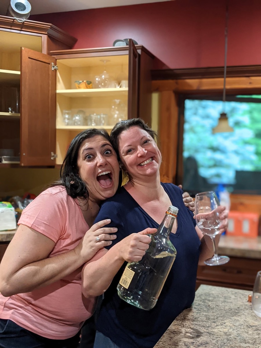 Two women in the cabin kitchen laughing at the camera, one holding a bottle of wine and a glass, red walls and a lit television behind.