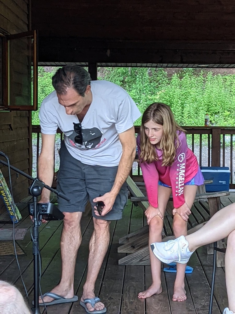A man bending over a fishing rod on the cabin deck while a girl in a pink top watches closely beside him.