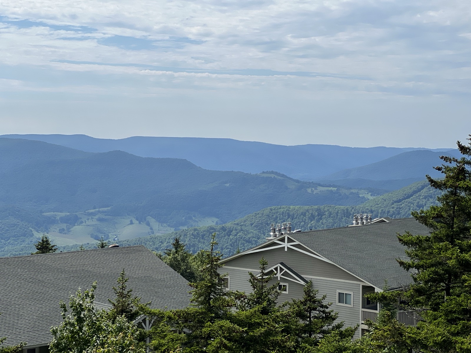 A view over the rooftops of Snowshoe village to layered blue mountain ridges under a soft cloudy sky, evergreens in the foreground.