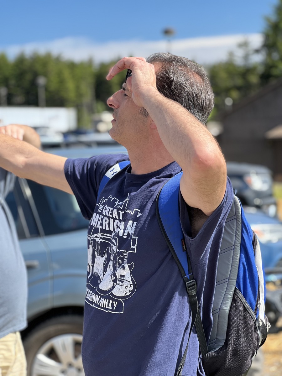 A man in a navy Great American Guitar Show t-shirt with a backpack, one hand raised to shade his eyes, looking upward in a parking lot.