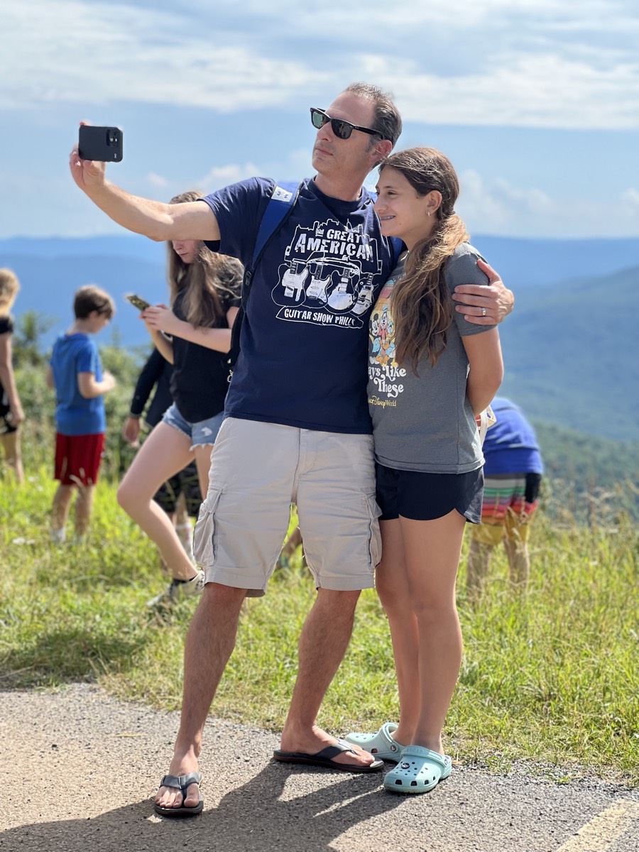 A man in sunglasses and a Great American Guitar Show t-shirt holding a phone up for a selfie with a girl tucked under his arm, a hazy mountain valley behind them.