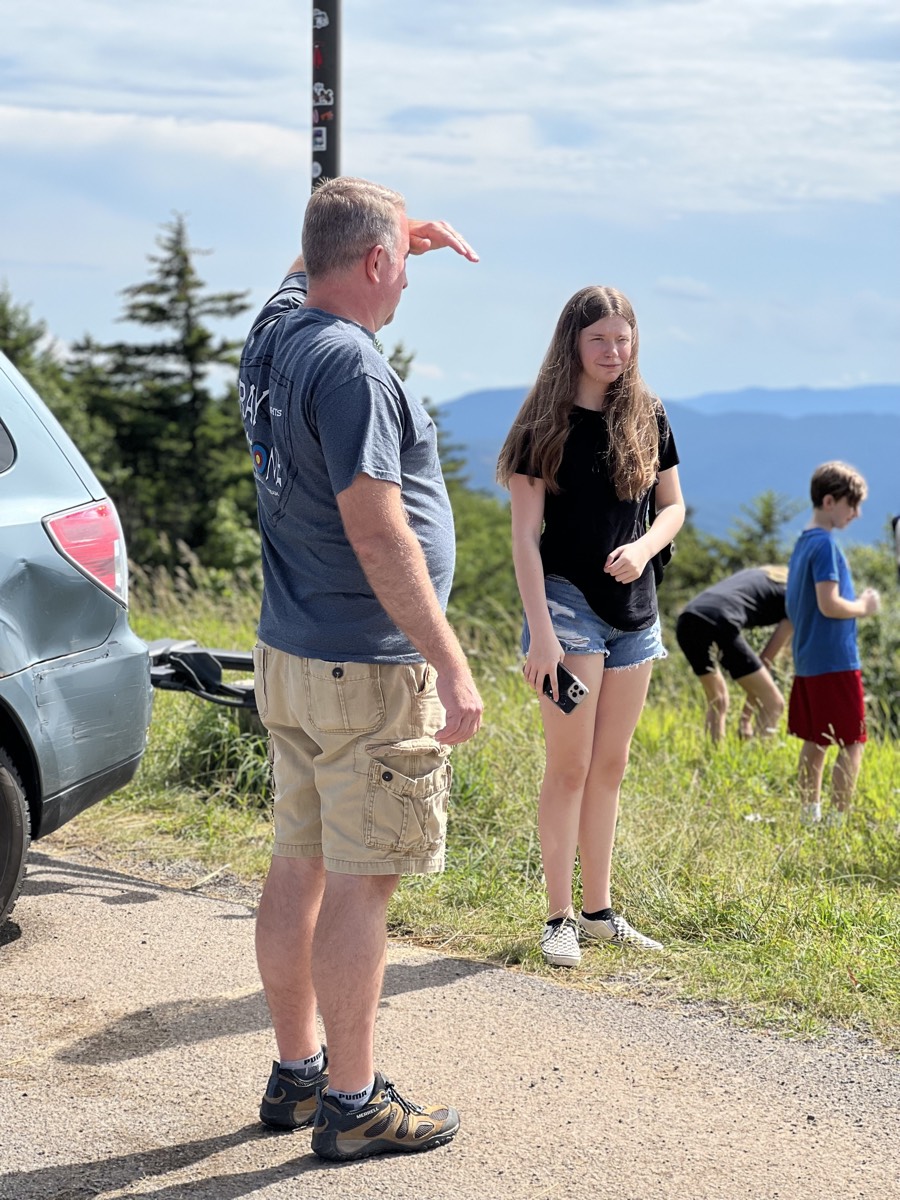 An older man in a blue shirt shielding his eyes from the sun while talking to a teenage girl at a roadside pull-off, mountains in the distance.