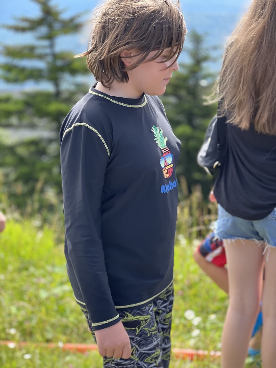 A boy in a black long-sleeved rash guard with a pineapple graphic standing in a grassy field, looking off to the side.