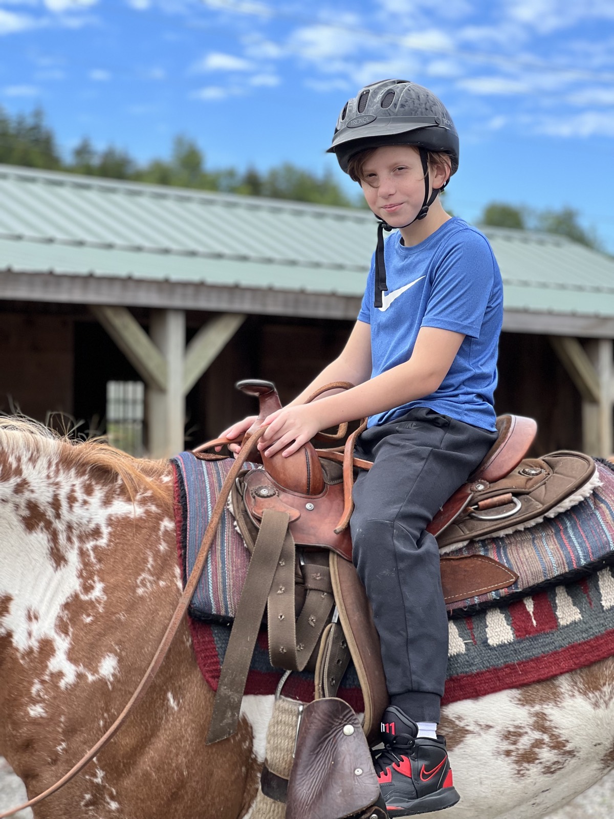 A boy in a blue shirt and riding helmet sitting on a brown and white paint horse in front of a stable, blue sky behind.