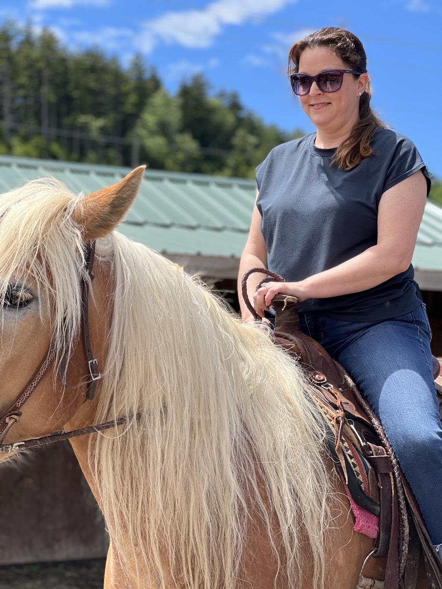 A woman in sunglasses and a dark t-shirt riding a palomino horse with a long pale mane, a stable roof behind her.