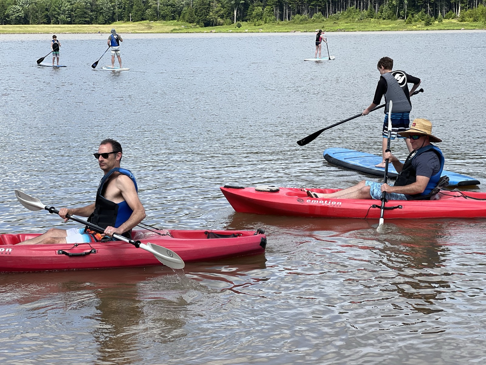 Two men in red kayaks and children on stand-up paddleboards spread across a calm lake, forested shoreline behind.