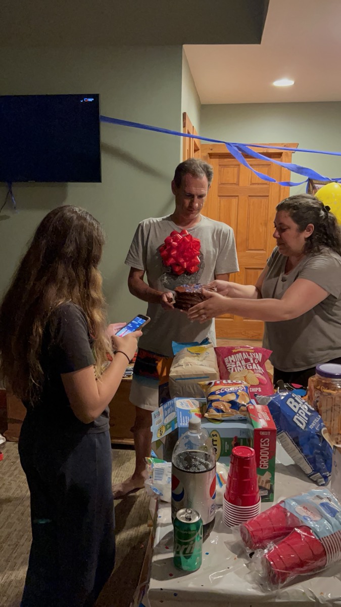 Lanay Zarallo handing a small cake topped with a large red bow to her husband Brian in a cabin living room, a table of chips, cups and drinks in the foreground.