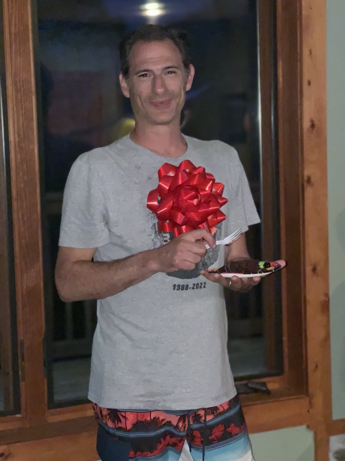 Brian Zarallo in a gray t-shirt printed with the dates 1988-2022, holding a small cake topped with a large red bow in one hand and a plate of chocolate cake with a fork in the other.