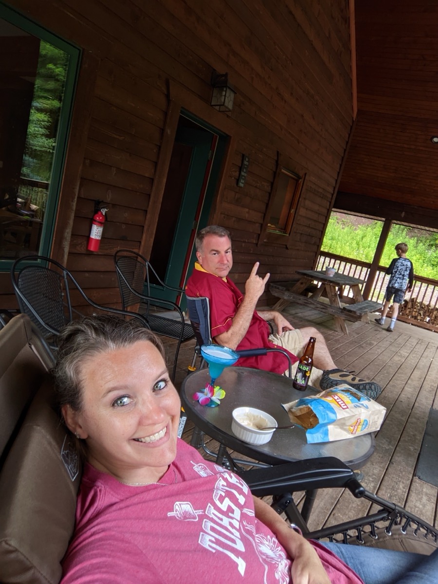 A woman smiling in a selfie from a porch recliner while a man in a maroon shirt throws a hand sign from his chair, drinks and snacks on the table between them.