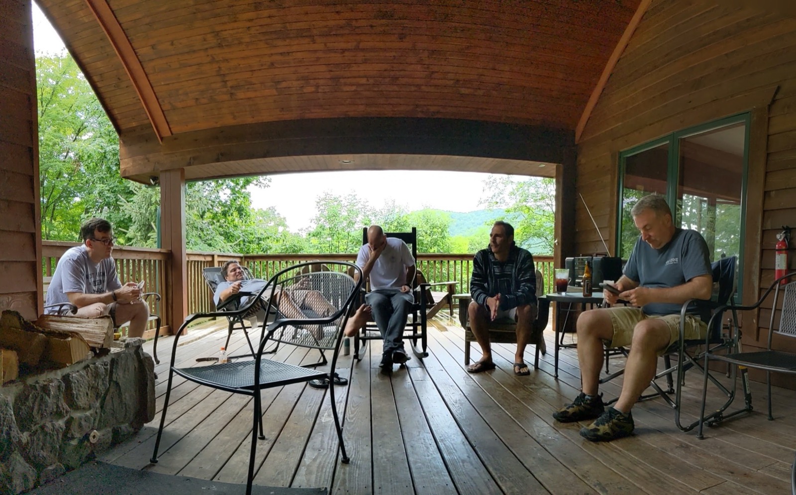 Five adults sitting in chairs spread across a wide covered cabin porch with a vaulted wooden ceiling, green trees and a mountain view beyond the railing.