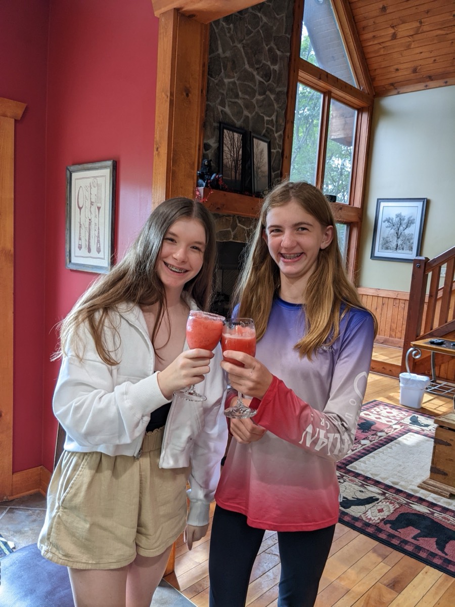 Two teenage girls grinning and touching tall glasses of frozen strawberry drinks together in a cabin great room with a stone fireplace behind them.