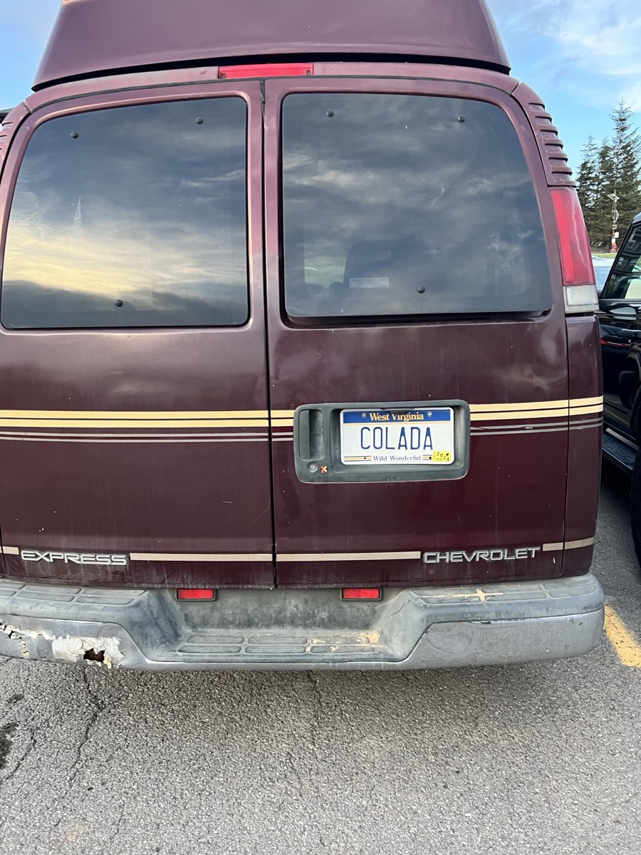 The back of a maroon Chevrolet Express conversion van in a parking lot, with a West Virginia license plate reading COLADA.