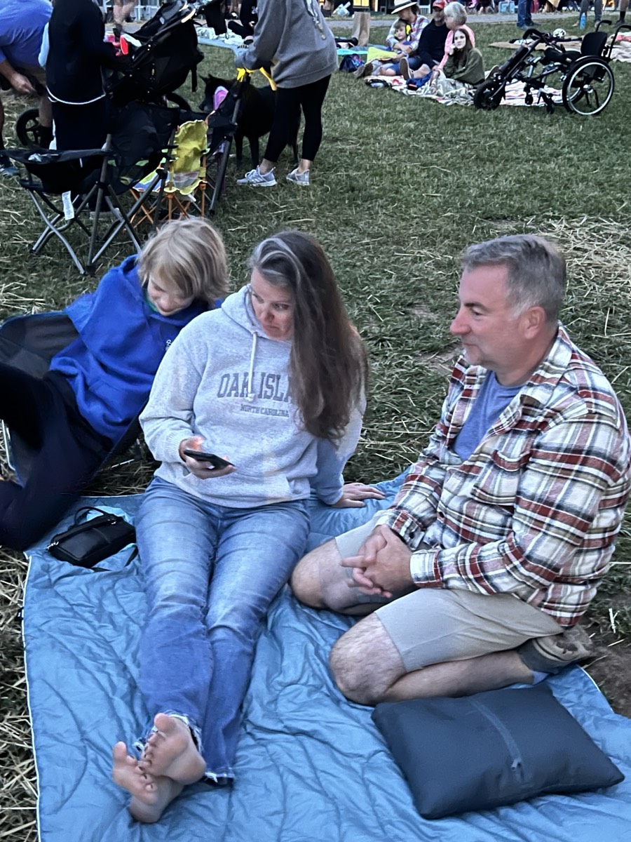 Two adults and a child sitting on a blue blanket on a grassy field at dusk, a crowd of people on blankets and camp chairs spread out behind them.