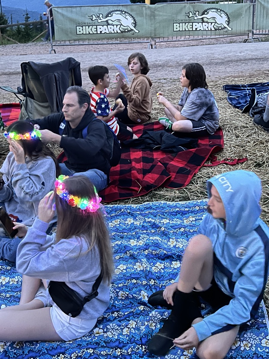 Children and adults sitting on plaid and floral blankets on straw in front of a Snowshoe Bike Park banner at dusk, several wearing glowing LED flower crowns.