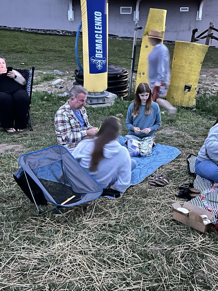 A man and two children sitting on a blue blanket on the grass beside a yellow and blue snow-making machine at dusk.