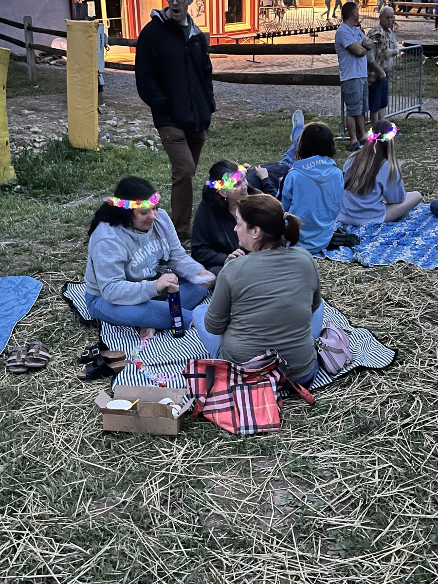 A group of women and children sitting on blankets on straw at dusk with drinks and a takeaway box between them, wearing glowing flower crowns.