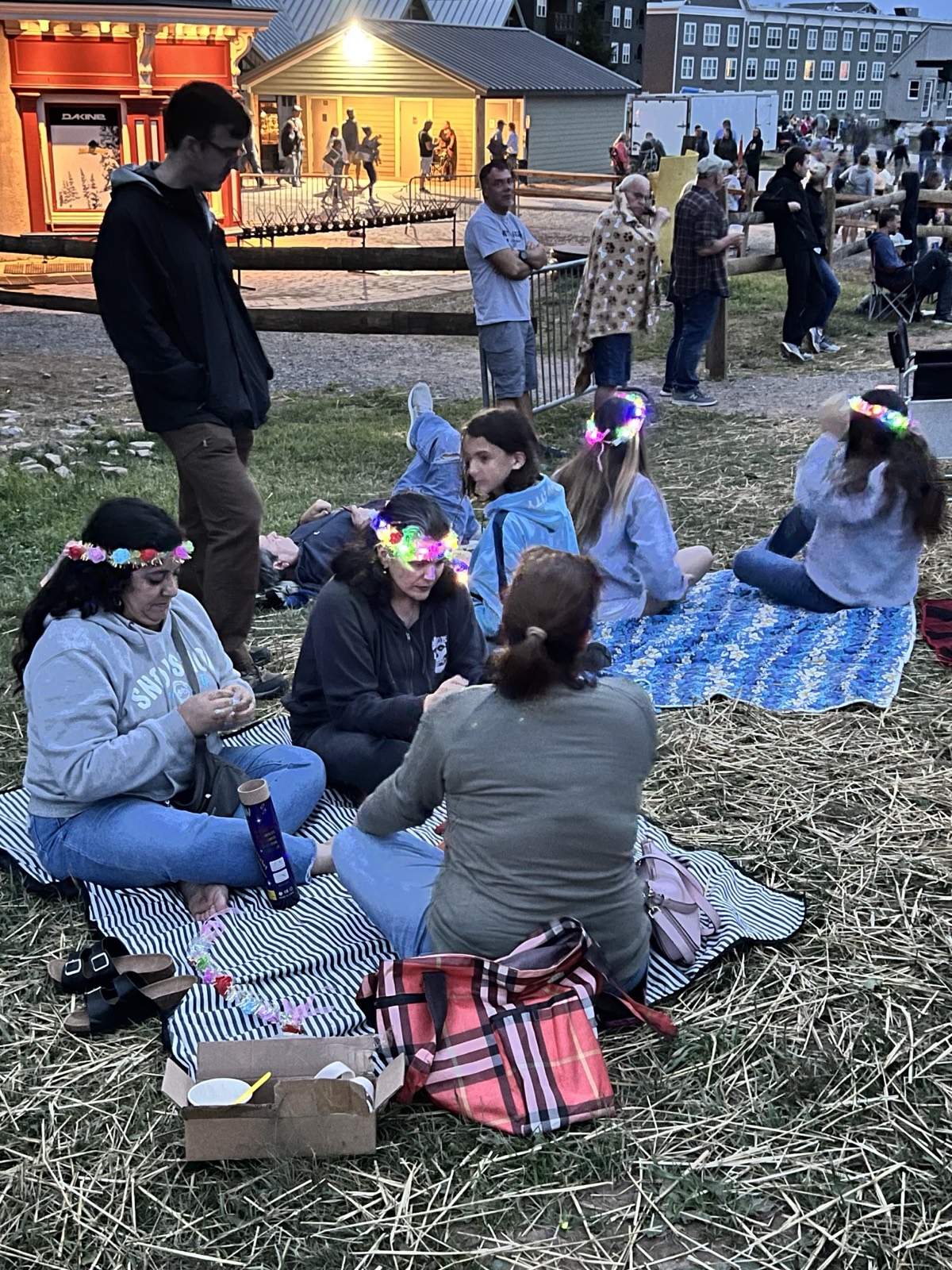 Families sitting on blankets spread over straw on a lawn at the top of Snowshoe mountain at dusk, light-up flower crowns on several heads, lit lodge buildings and people milling about behind them, all waiting for the Fourth of July fireworks.