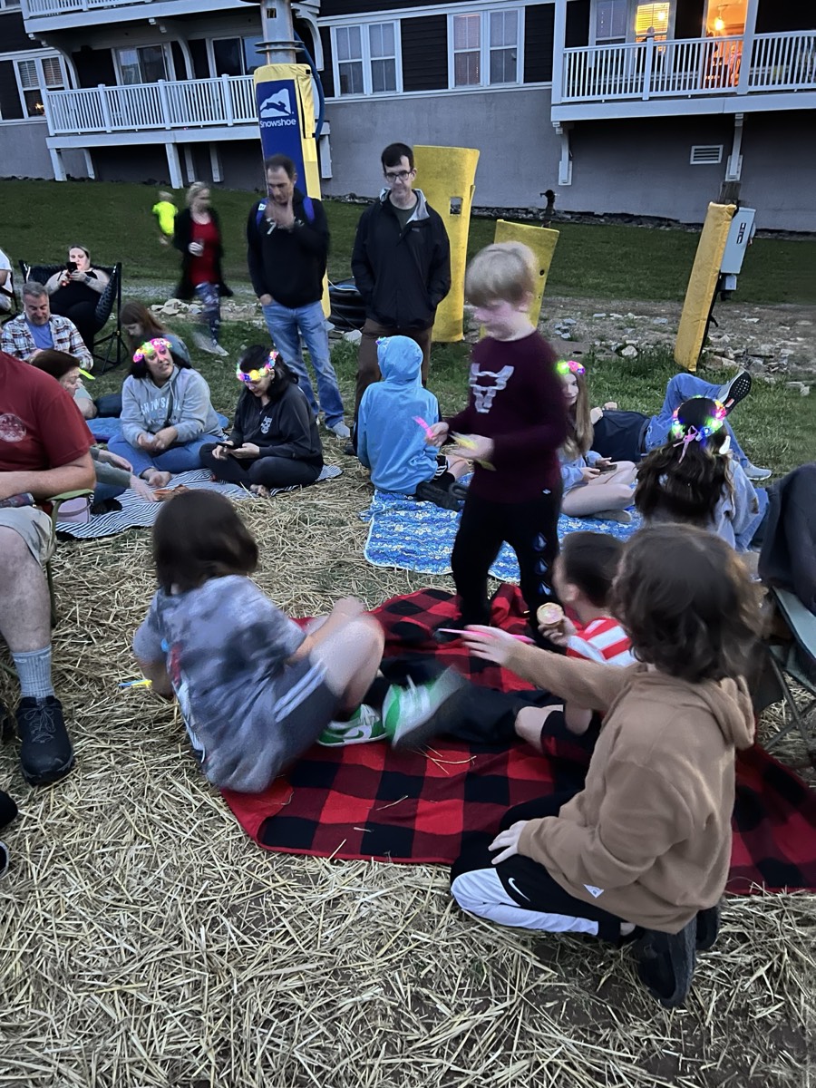 Children sitting and standing on a red plaid blanket on straw at dusk, adults in camp chairs behind them and a lit building beyond.