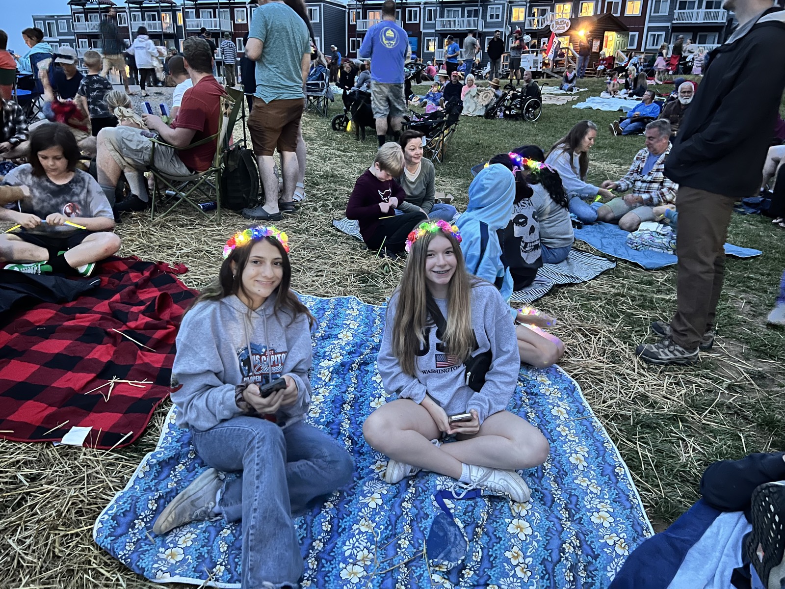 Two teenage girls in glowing flower crowns sitting cross-legged on a blue floral blanket at dusk, smiling at the camera, a crowd on the lawn behind them.