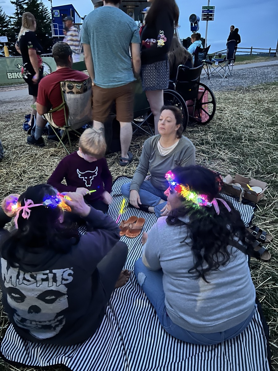 Adults and children sitting on a striped blanket on straw at dusk wearing light-up flower crowns, a bike park sign and camp chairs behind them.