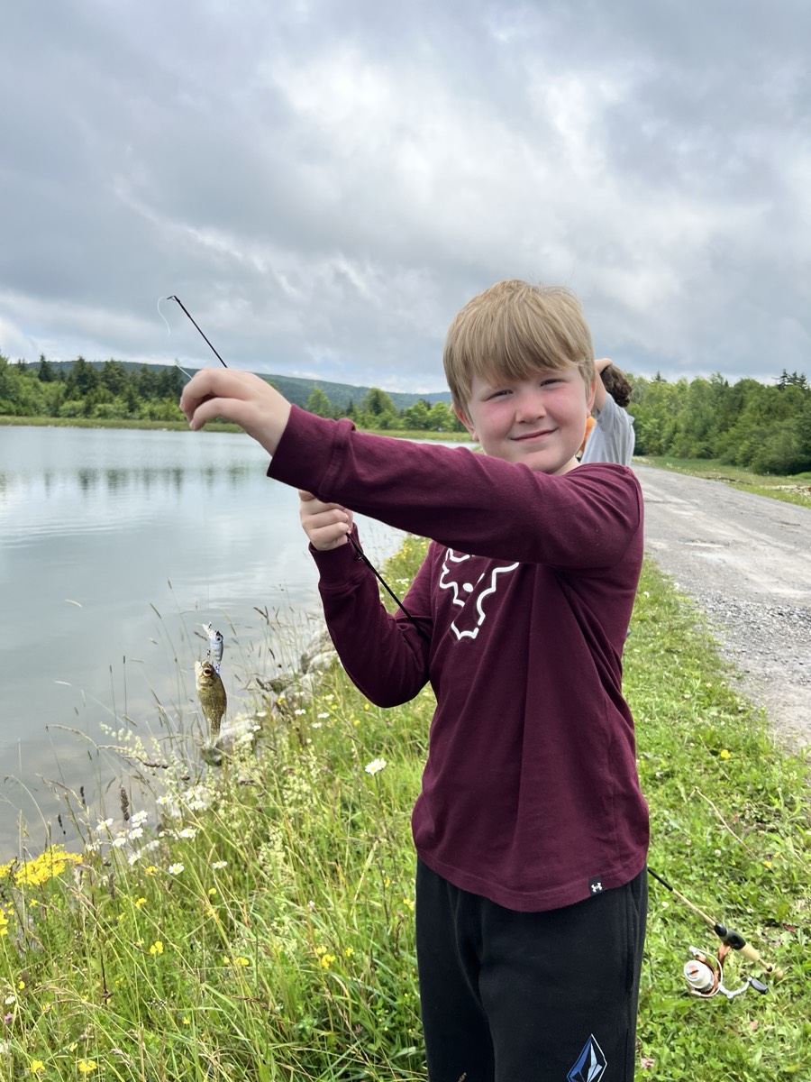 A blond boy in a maroon long-sleeved shirt smiling and holding his line up so the small fish on the end swings clear of the grass.