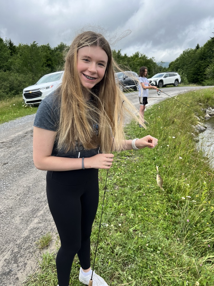 A girl with long blonde hair blowing across her face, smiling and holding a fishing rod with a small fish on the line, cars parked along the gravel road behind.
