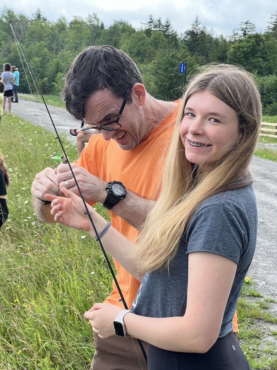 A man in an orange t-shirt and glasses working on a fishing line while a smiling girl with braces holds the rod steady beside him.