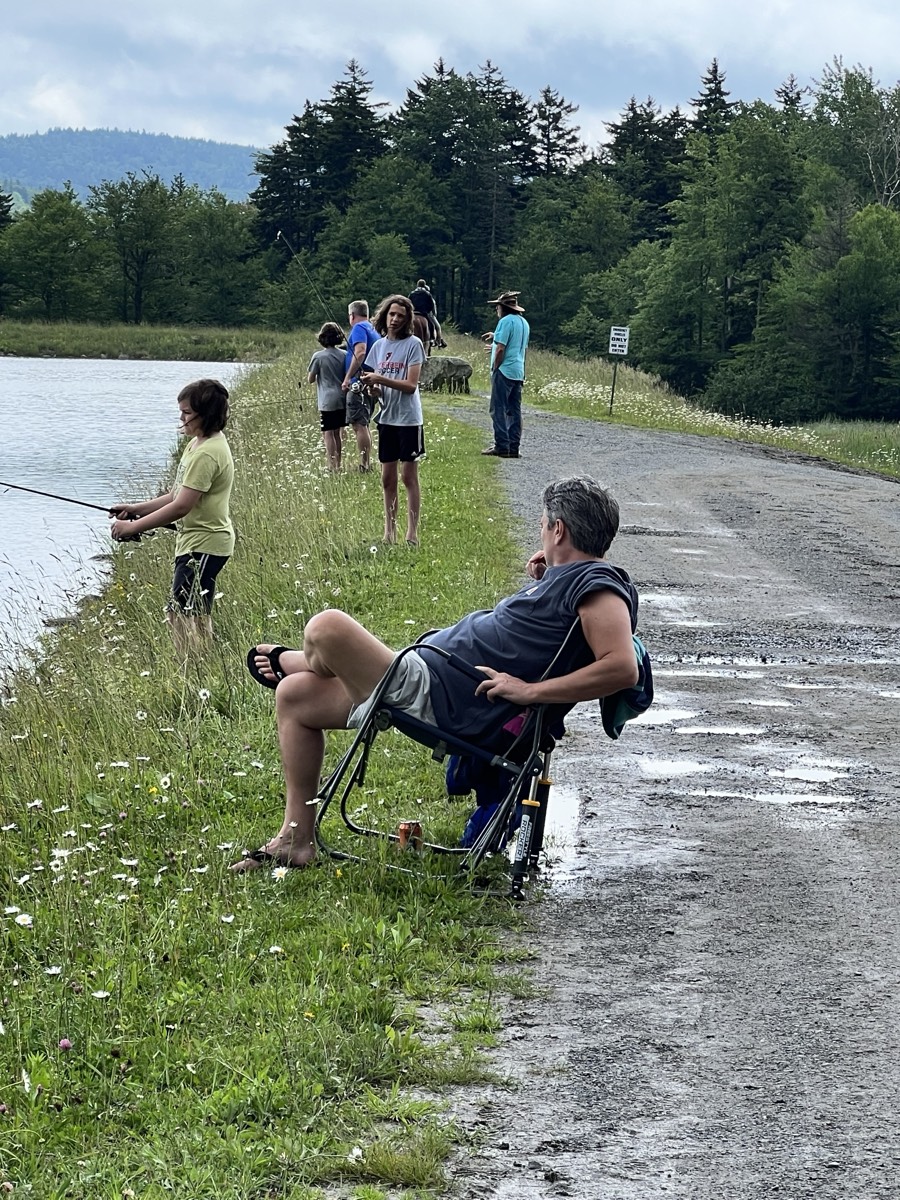 A man in an orange t-shirt and a girl in a gray top standing at the edge of the pond with their backs to the camera, working a fishing rod together.