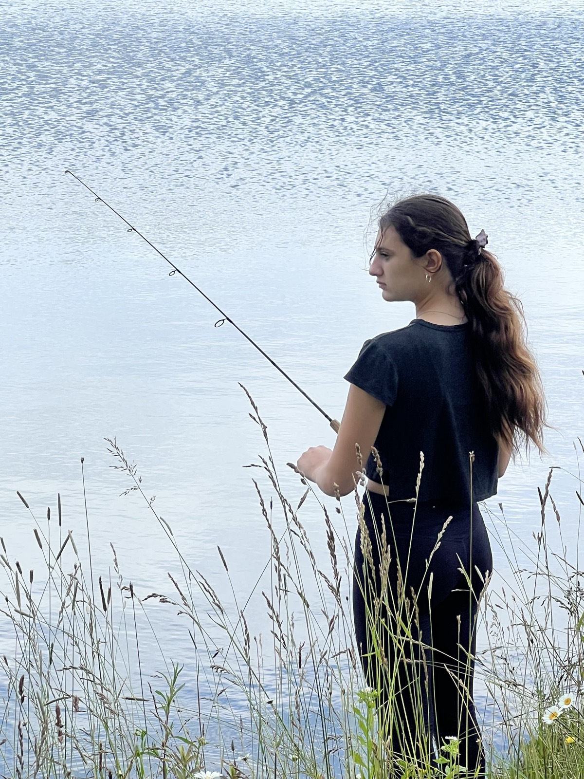 A girl in a dark top standing in tall grass at the edge of a still pond, fishing rod out over the water, seen from behind.