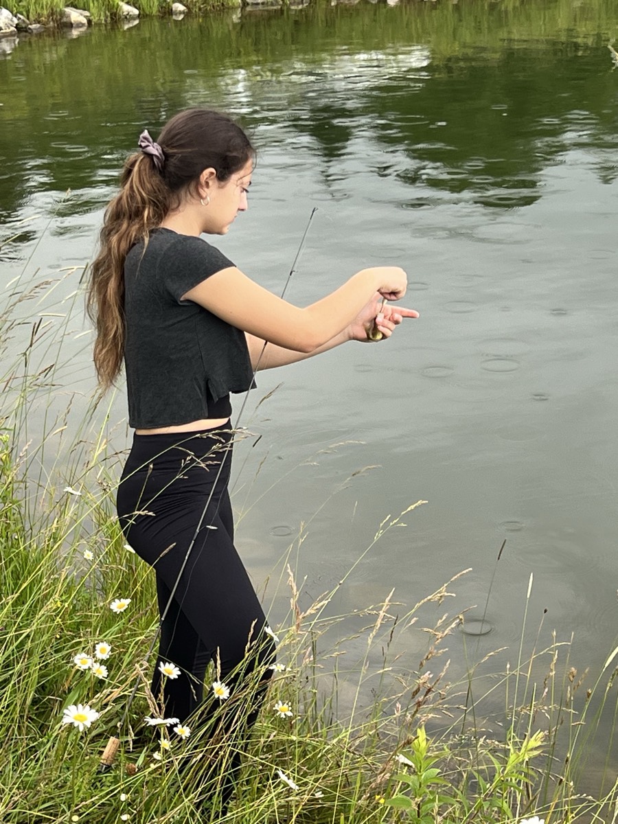 The same girl leaning over the water at the pond edge, carefully holding a small fish in both hands to release it.