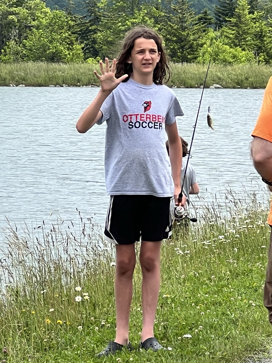 A child in an Otterbein Soccer t-shirt holding up five fingers and grinning by the pond, another child fishing in the background.